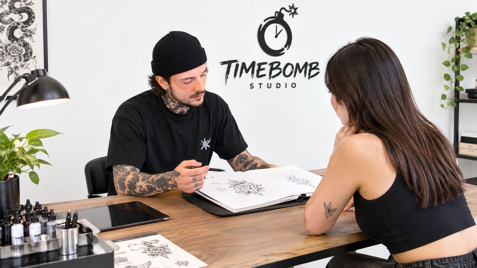A professional tattoo artist in a studio discusses a portfolio design with a female client at a desk.