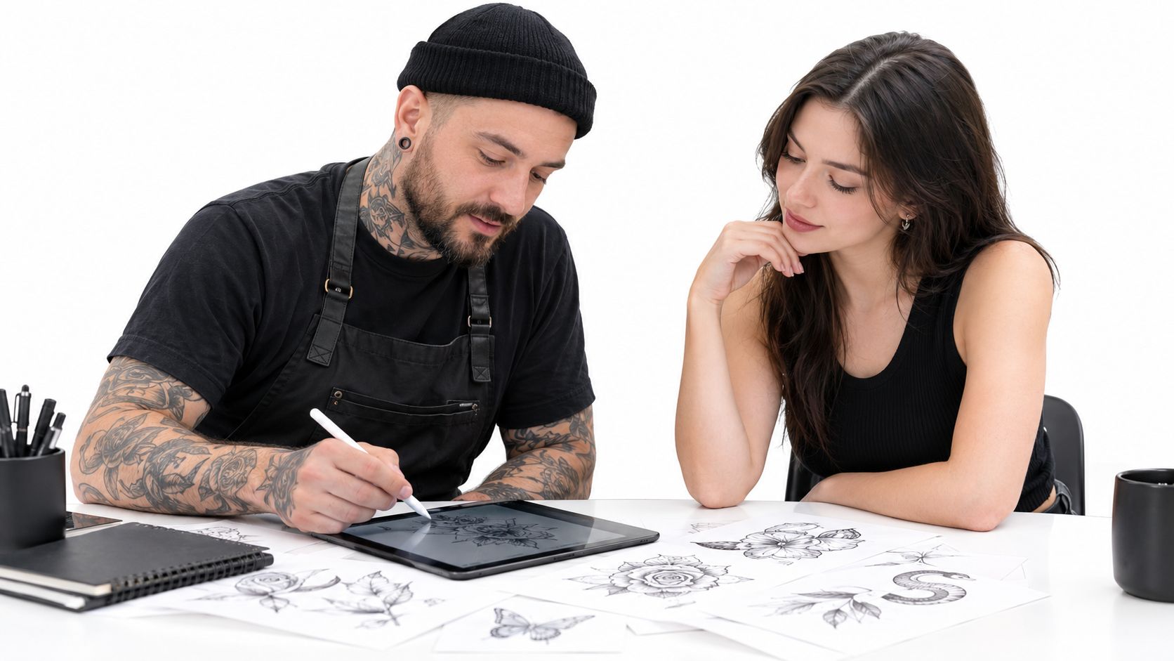 A professional tattoo artist in an apron discussing tattoo designs on a digital tablet with a female client.